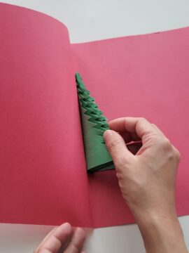 Person gluing paper Christmas tree to crease of a folded piece of red construction paper