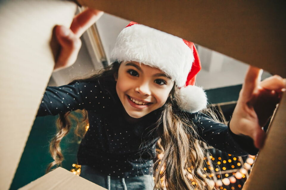 https://www.littlepassports.com/wp-content/uploads/2022/10/girl-wearing-Santa-hat-smiling-and-looking-inside-a-box-960x640.jpg